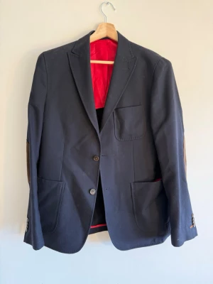 Morris Stockholm - Navy kavaj - Morris Stockholm – marinblå kavaj med bruna mocka-armbågslapper och helrött foder med röd piping. Patch-fickor, två knappar, notch lapel. Karaktärsfull piece med bra detaljer. Stl 50. Skick: mycket bra