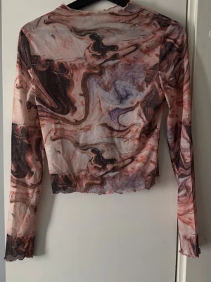 Mesh topp med marmormönster från SHEIN - Trendig mesh topp från SHEIN med lång ärm och marmorinspirerat mönster i rosa, lila och bruna toner. Toppens material är tunn och lätt, perfekt för lager-på-lager. Avslutet nertill och vid ärmslut har en vågig kant för extra detalj.