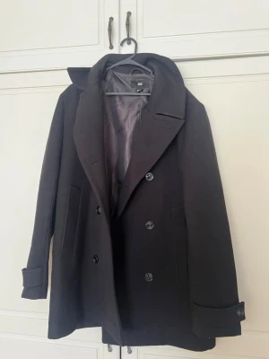 Svart skepparkavaj från H&M - Stilren svart skepparkavaj från H&M med dubbelknäppning, breda slag och två sidofickor. Kavajen har klassisk passform, slät yta och är helfodrad för extra komfort. Perfekt för dig som gillar en tidlös look.