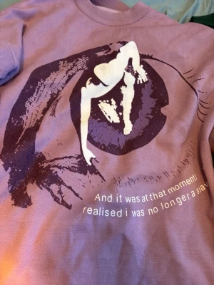 Lila sweatshirt med konstnärligt tryck - Cool lila sweatshirt med ett stort konstnärligt tryck i vitt och mörklila på framsidan. Trycket föreställer en stiliserad figur och texten 'And it was at that moment I realised I was no longer a slave'. Rund hals och avslappnad passform.