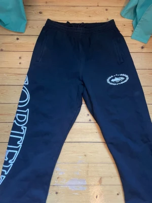 Svarta mjukisbyxor Corteiz - Svarta mjukisbyxor från Corteiz med vit tryckt logga på ena benet och emblem på höften. Byxorna har resår i midjan och två sidofickor. Tillverkade i mjukt material som passar perfekt för chill eller träning.