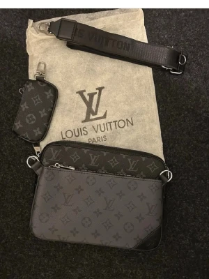  axelväska monogram svart - Snygg svart axelväska  med klassiskt monogrammönster i grått och svart. Väskan har justerbar axelrem och en mindre matchande pouch. Tillverkad i canvas med detaljer i läder och metall. Perfekt för dig som gillar exklusiva accessoarer.