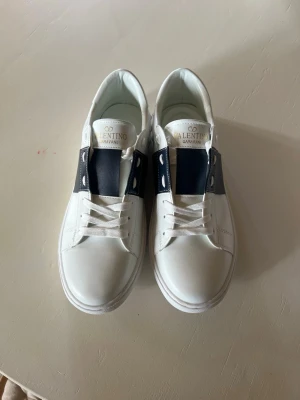 Valentino Garavani vita sneakers - Snygga vita sneakers från Valentino Garavani med bred mörkblå rem över vristen och klassiska vita snören. Skorna har en stilren design i läder och låg profil som passar till många olika outfits. Dessa skor passar bra 44 men även 43