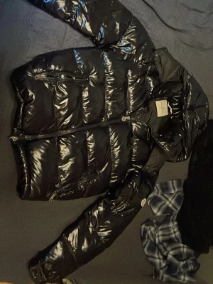 Svart glansig pufferjacka Moncler - Säljer en svart glansig pufferjacka från Moncler med hög krage och dragkedja framtill. Jackan har två snedställda fickor med dragkedja och klassisk Moncler-logga på ärmen. Perfekt för kalla vinterdagar och riktigt snygg streetstil.