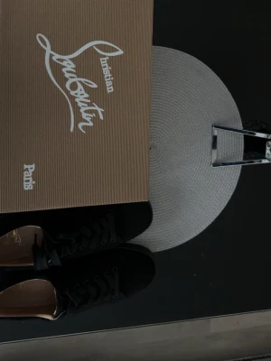 Christian Louboutin svarta sneakers - Stilrena svarta sneakers från Christian Louboutin med ikonisk röd sula och präglad logga undertill. Skorna har lågt skaft, svarta skosnören och ovandel i läder för en lyxig känsla. Perfekt för dig som gillar exklusiva detaljer och tidlös design.