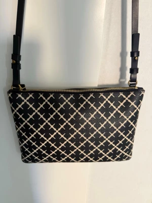 Malene Birger väska - Snygg svart axelväska med vitt grafiskt rutmönster och justerbar axelrem. Väskan har guldfärgade metalldetaljer och dragkedja upptill. Perfekt för dig som gillar stilrena accessoarer med en twist. Materialet ser ut att vara konstläder. Använd men ser ut som ny och är fräsch inuti