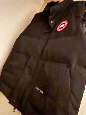 Svart dunväst från Canada Goose - Säljer en svart dunväst från Canada Goose med klassisk logotyp på bröstet. Västen har hög krage, tryckknappar framtill och två stora fickor. Perfekt för lager-på-lager och riktigt varm tack vare dunfyllningen.