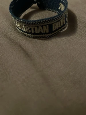 Blått Christian Dior J'Adior armband, - Snyggt blått tygarmband från Christian Dior med texten 'J'Adior' broderad i vitt. Armbandet har justerbara knytband och dekorativa detaljer längs kanterna. Perfekt accessoar för att lyfta din stil.