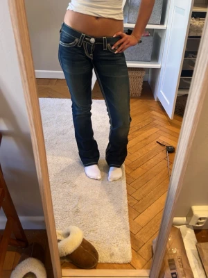 Vintage True Religion lågmidjade bootcut jeans mörkblå  - Säljer ett par mörkblå bootcut jeans från True Religion med tydliga vita kontrastsömmar och klassiska fickdetaljer bak. Jeansen har låg midja och är tillverkade i denim med en snygg tvätt. Jeansen är vintage men är i extremt bra skick och känns som nya och tyget är fortfarande tjockt. MÅTTEN ÄR midja: ca 36-40cm, innebens längden är ca 75cm 