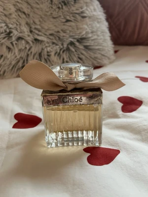 Chloé Eau de Parfum med beige rosett - Elegant parfym från Chloé i en klassisk, rektangulär glasflaska med räfflad design. Flaskan har en silverfärgad metallkork och dekoreras med en beige tygrosett runt halsen. Doften är feminin och tidlös, perfekt för dig som gillar lyxiga parfymer.
