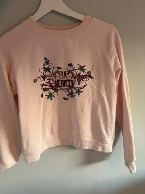 Ljusrosa  Zadig Voltaire sweatshirt med tryck - Ljusrosa sweatshirt från Zadig Voltaire  med coolt tryck framtill i svart och rött med blommor och text. Tröjan har rund halsringning och långa ärmar. Perfekt för dig som gillar streetstyle och vill ha något unikt i garderoben.