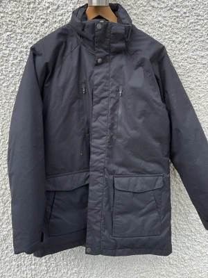 Svart parkas från Everest - Stilren svart parkas från Everest med hög krage och dragkedja samt tryckknappar framtill. Jackan har två stora fickor med lock och två bröstfickor med dragkedja. Tillverkad i slitstarkt material, perfekt för kalla dagar.