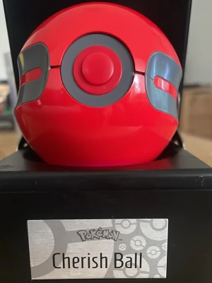 Pokémon Cherish Ball med LED-ljus - Snygg Cherish Ball från Pokémon i röd och grå plast med rund form. Bollen har inbyggd LED-belysning som lyser upp när den aktiveras. Perfekt som samlarpryl eller dekoration för Pokémonfans. Levereras i en svart displaybox med metallskylt.