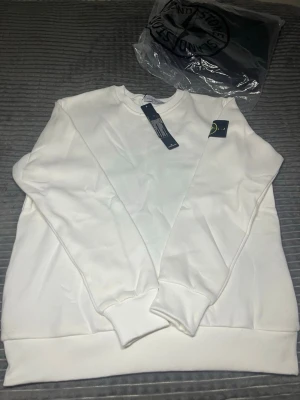Vit sweatshirt från Stone Island - Vit sweatshirt från Stone Island med klassisk rund hals och ribbade muddar. Tröjan har den ikoniska Stone Island-loggan på vänster ärm. Perfekt för en clean och stilren look.