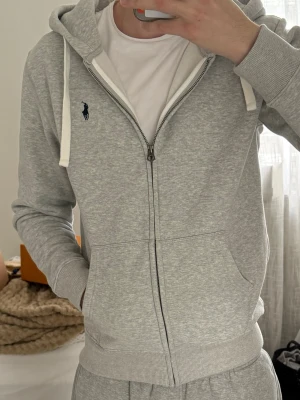 Grå zip-hoodie från Polo Ralph Lauren - Snygg ljusgrå hoodie med dragkedja från Polo Ralph Lauren. Klassisk design med vit dragsko i huvan, två fickor fram och broderad marinblå logga på bröstet. Mjuk bomullsblandning och ribbade muddar för extra komfort.