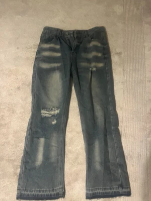 Blå slitna raka jeans - Säljer ett par blå jeans med raka ben och slitna detaljer på framsidan. Jeansen har en tvättad look med ljusare partier och klassiska fem fickor. Perfekta för en avslappnad och trendig stil.