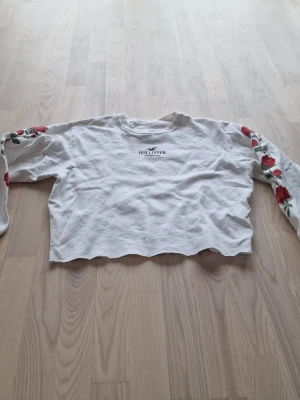 Vit cropped hoodie från Hollister - Vit cropped hoodie från Hollister med broderade röda rosor längs ärmarna och logga på bröstet. Tröjan har långa ärmar och rå kant nertill för en trendig look. Perfekt för dig som gillar en avslappnad och cool stil.