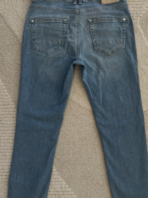 Snygga ljusblå jeans från Mos Mosh - Snygga blå jeans från Mos Mosh med klassisk femficksdesign och gulddetaljer vid fickorna framtill. Jeansen har en rak passform och är tillverkade i mjukt denimtyg. Perfekta för en avslappnad och trendig look.