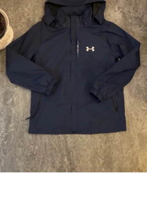 Blå vindjacka från Under Armour - Snygg marinblå vindjacka från Under Armour med huva, dragkedja och två sidofickor med dragkedja. Jackan har elastiska muddar vid ärmsluten och meshfoder på insidan för extra komfort. Perfekt för blåsiga dagar och sportiga outfits.