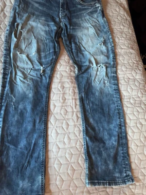 Blå slitna jeans från Evisu - Säljer ett par blå jeans från Evisu med slitningar och hål på framsidan för en riktigt trendig och avslappnad look. Jeansen har raka ben och klassisk femficksmodell. Perfekta för dig som gillar streetwear och vill sticka ut lite extra. w34 L34