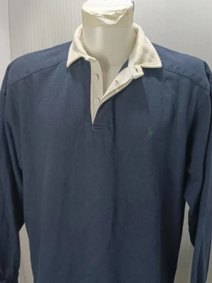 Blå långärmad piké från Ralph Lauren - Klassisk blå långärmad piké från Ralph Lauren med vit krage och knäppning framtill. Tröjan har lång ärm och diskret grön logga broderad på bröstet. Tillverkad i mjuk bomull som ger en avslappnad och sportig look.