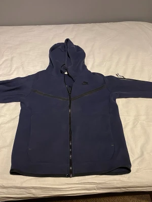 Mörkblå Nike hoodie med dragkedja - Snygg mörkblå hoodie från Nike med hel dragkedja framtill och två stora fickor. Klassisk huva och diskret Nike-logga på bröstet. Perfekt för en avslappnad streetstyle-look. Tillverkad i mjukt material för extra komfort.