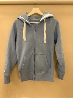 Ljusblå hoodie från Russemerch - Ljusblå hoodie med dragkedja från Russell Athletic. Tröjan har vita snören i huvan, två stora fickor framtill och ribbade muddar vid ärmslut och nederkant. Broderad logga på ena ärmen. Perfekt för en avslappnad stil.