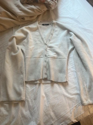 Beige cropped kofta från Gina Tricot - Säljer en beige cropped kofta från Gina Tricot med v-ringning och ribbad midja. Koftan har långa ärmar och knappar framtill. Perfekt för lager på lager och enkel att styla till olika outfits.