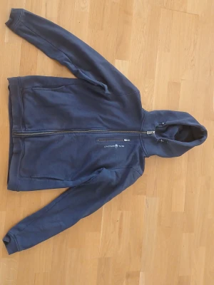 Mörkblå hoodie Sail Racing - Mörkblå hoodie med hel dragkedja och huva från Sail Racing. Tröjan har en bröstficka med dragkedja och logotyp på framsidan. Långa ärmar och klassisk passform, perfekt för dig som gillar en enkel och stilren look. Den har storlek 176, S eller 46