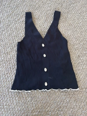 Svart ribbad linnetopp med snäckor - Cool svart ribbad linnetopp med breda axelband och v-ringning. Framsidan har dekorativa vita knappar och nederkanten är vågig med vit kant. Perfekt för att skapa en avslappnad och trendig look.