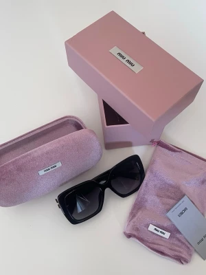 Svarta solglasögon från Miu Miu - Stilrena svarta solglasögon från Miu Miu med stora, fyrkantiga bågar. Kommer med lyxigt rosa fodral och matchande tygpåse. Perfekta för dig som vill sticka ut med en trendig accessoar från ett ikoniskt märke.
