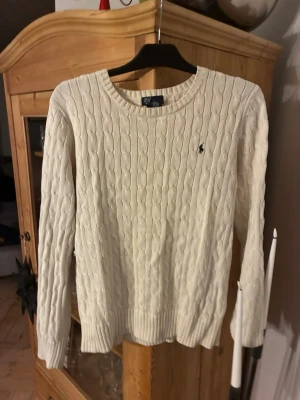Vit kabelstickad tröja Polo Ralph Lauren - Kabelstickad vit tröja från Polo Ralph Lauren i 100% bomull. Klassisk rund halsringning och broderad logga på bröstet. Perfekt för en stilren och avslappnad look med långa ärmar och ribbade muddar.