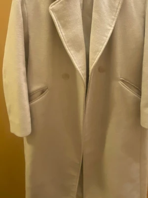 Beige ullrock från H&M - Stilren beige ullrock från H&M med klassisk krage och två knappar framtill. Rocken har två snedställda fickor och en rak, längre passform som ger en snygg siluett. Perfekt för dig som gillar minimalistisk och tidlös stil.