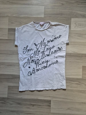 Vit t-shirt med svart textprint - Vit t-shirt med coolt svart textprint över bröstet där olika städer står skrivna i snirklig stil. Klassisk passform med korta ärmar och rund halsringning. Perfekt för en avslappnad och trendig look. Den har 3 små hål fram och ett bak på vänster axel.