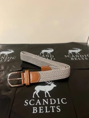 Grått flätat bälte  - Stilrent grått flätat bälte med detaljer i brunt skinn och silverspänne. Perfekt accessoar för att lyfta din outfit med en modern touch. Bältet har en flexibel och bekväm design. 105cm