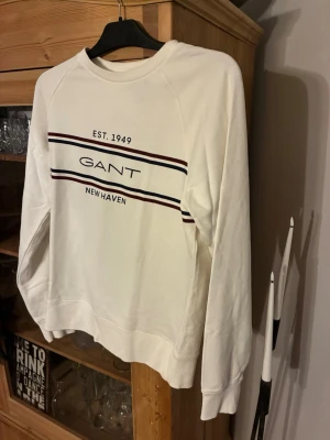 Vit sweatshirt från Gant - Vit sweatshirt från Gant med tryck på bröstet: 'EST. 1949 GANT NEW HAVEN' och två ränder i marinblått och vinrött. Tröjan har rund hals, långa ärmar och ribbade muddar. Perfekt för en avslappnad och stilren look. Använd ett fåtal gånger och har storleken M