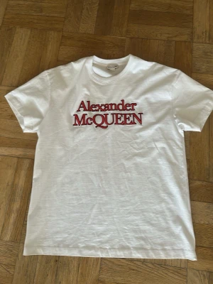 Vit Alexander McQueen t-shirt - Säljer en klassisk vit t-shirt från Alexander McQueen med röd logotyp framtill. T-shirten har rund hals och korta ärmar, perfekt för en clean och stilren look. Tillverkad i mjuk bomull för maximal komfort.