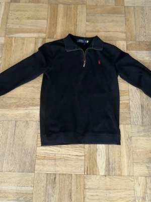 Svart halvzip tröja från Polo Ralph Lauren - Klassisk svart tröja med halv dragkedja från Polo Ralph Lauren. Tröjan har en liten röd broderad logga på bröstet och ribbade muddar vid ärmslut och nederkant. Perfekt för en stilren och avslappnad look.