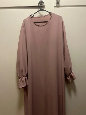 Ljusrosa abaya med volangärm - En stilren ljusrosa abaya med rund halsringning och långa ärmar med volangdetalj vid ärmslut. Abayan har en rak passform och är tillverkad i ett mjukt, slätt material som ger ett elegant intryck.💓 