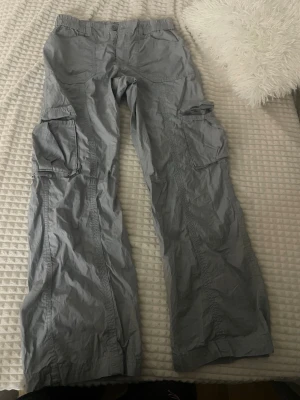Grå cargobyxor från Gina Tricot - Säljer ett par grå cargobyxor från Gina Tricot med breda ben och stora sidofickor. Byxorna har resår i midjan och är tillverkade i ett lätt, lite prassligt material. Perfekta för en avslappnad och trendig look.