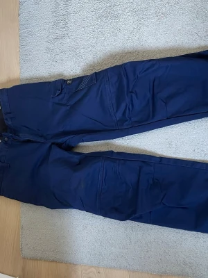 Mörkblå cargobyxor/arbetsbyxor med fickor  - Snygga mörkblå cargobyxor med flera praktiska fickor på benen och klassisk cargomodell. Byxorna har raka ben och är tillverkade i ett slitstarkt bomullsmaterial. Perfekta för dig som gillar en avslappnad och funktionell stil. Perfekt för dig som behöver ett par slitstarka byxor att kunna jobba i när du till exempel donar med bilen. Byxorna har en oljefläck på ena benet . 