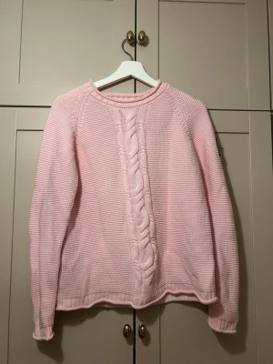 ljusrosa stickad tröja med kabelmönster - Ljusrosa stickad tröja från Bondelid med klassiskt kabelmönster framtill och rund halsringning. Tröjan har lång ärm och en patch med amerikansk flagga och B på vänster ärm. Perfekt för dig som gillar en söt och avslappnad stil