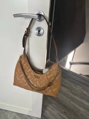 Senapsgul/Beige quiltad handväska från Guess - Snygg senapgul/ beige handväska från Guess med quiltat mönster och guldfärgad kedjedetalj. Väskan har en justerbar axelrem i brunt och Guess-logga framtill. Tillverkad i konstläder och perfekt för dig som vill ha en trendig accessoar.