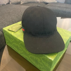 Svart keps från Gucci - Snygg svart keps från Gucci med diskret mönster och röd-gröna detaljer på sidan. Kepsen har en justerbar rem baktill för perfekt passform. Perfekt för att ge din outfit en lyxig touch.