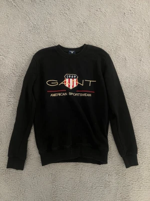 Svart sweatshirt från GANT - Svart sweatshirt från GANT med tryckt logga och texten 'American Sportswear' framtill. Rund halsringning och ribbade muddar vid ärmslut och nederkant. Klassisk och stilren design med broderad sköld i rött och vitt. Kanske lite liten i storlek