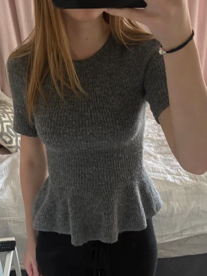 Grå ribbad topp med volangkant - Säljer en grå ribbad topp med korta ärmar och volangkant nedtill. Toppen har en rund halsringning och sitter figurnära upptill med en lösare passform nedtill. Perfekt för dig som gillar en enkel men ändå söt stil.