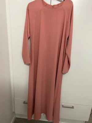 Lång rosa abaya  - En enkel och stilren långklänning i mjukt rosa tyg. Klänningen har långa ärmar och rund halsringning, perfekt för dig som gillar minimalistisk och avslappnad stil. Kommer i perfekt skick utan skador. Ganska mjuk material som passar perfekt för sommaren 