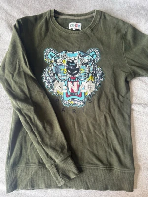 Olivgrön Kenzo sweatshirt  - Säljer en olivgrön sweatshirt från Kenzo med det ikoniska tigerhuvudet broderat i blått, vitt, gult och svart på bröstet. Tröjan har rund halsringning, långa ärmar och ribbade muddar. Perfekt för dig som vill sticka ut med en streetstyle-look.