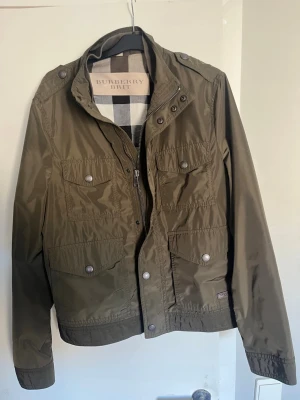 Olivgrön vindjacka från Burberry Brit - Stilren olivgrön vindjacka från Burberry Brit med fyra framfickor med lock och tryckknappar, hög krage och dragkedja med knappslå. Jackan har klassiskt rutigt foder och är tillverkad i ett lätt, vattenavvisande material. Perfekt för en trendig och avslappnad look.