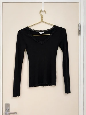 Svart långärmad topp med spets - Snygg svart långärmad topp med v-ringning och spetsdetaljer runt halsen. Modellen är slim fit och har ribbad struktur samt vågig kant vid ärmslut och nederkant. Perfekt att styla med jeans eller kjol för en trendig look.